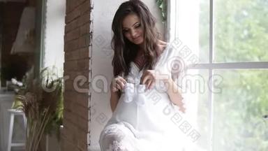 怀孕的，漂亮的女人坐在窗边的窗台上。 抱着婴儿`鞋袜，在她身上踩上几步